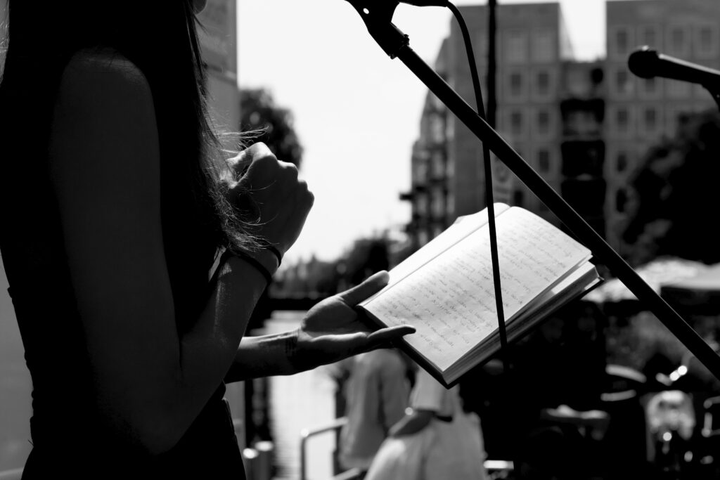 Zwart-wit foto van mijn notitieboek, dat ik vasthoud in mijn linkerhand, terwijl ik achter de microfoon sta op een podium. Mijn rechterhand is in een ontspannen vuist gebald. Je kunt net niet lezen wat er op de pagina's staat.